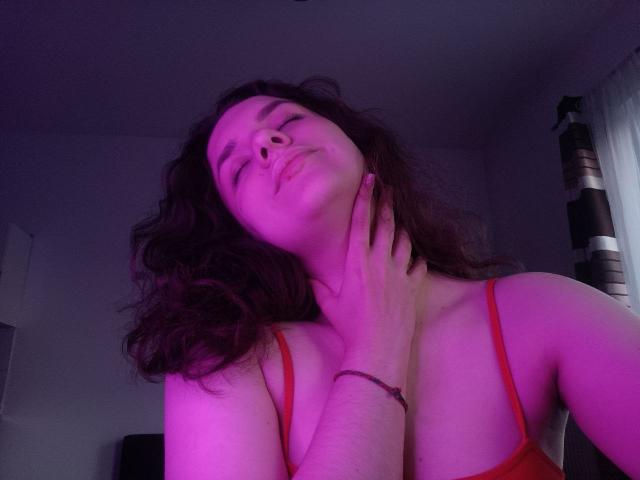 Dreia with curly dark hair, eyes closed, red tank top, hand on neck, purple lighting, slightly tilted head, soft expression, visible cleavage, red bracelet, subtle smile.
