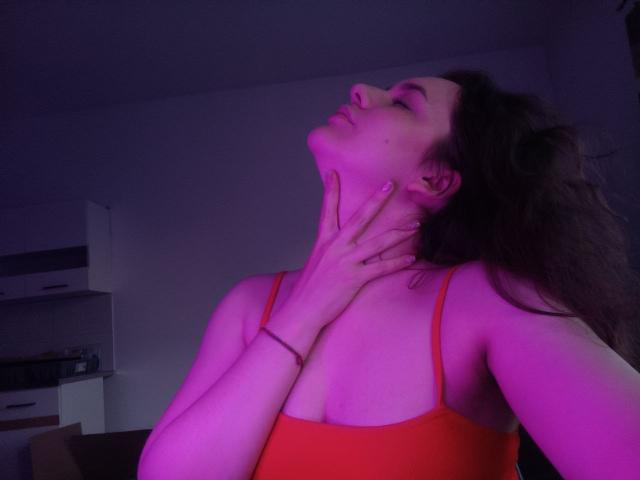 Dreia with purple-lit skin, eyes closed, head tilted back, hand on neck, wearing a red tank top, exposing her tits, in a dim room.