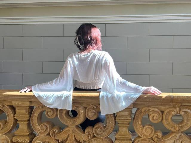 Meeduza with red hair, back to camera, wearing white flowy blouse, black waistband, standing on ornate wooden balcony, sunlit, hands on railing.