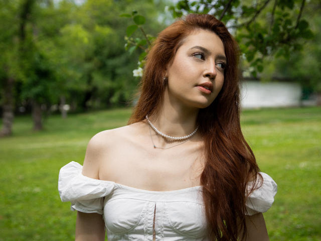 EllyCat has long red hair, white off-shoulder dress, pearl necklace, looking up with a slight smile, grassy outdoor background, soft light, her tits are visible.