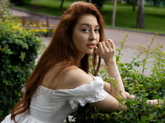 EllyCat with long red hair, off-shoulder white dress, and freckles, resting her chin on her hand, looking at the camera, in a green garden with bushes.