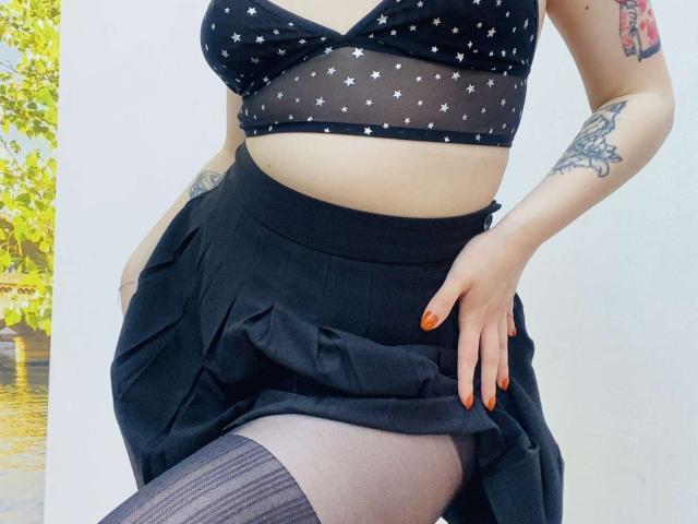 KarKutie with her tits showing in a sheer black star-patterned top, black skirt, and gray thigh-highs, hand on hip, orange nails, tattoos on arm, outside in daylight.