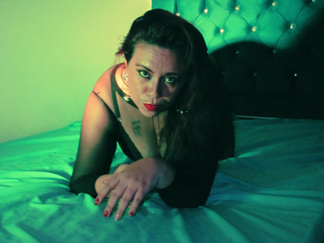 LexyDom is on a bed, wearing black lingerie, with her tits visible, leaning forward, green and yellow lighting, red lipstick, black hair, and a tattoo on her chest.
