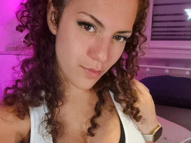 OliviaOcean-hot with curly brown hair, brown eyes, and a small smile, wearing a white sleeveless top, showing her tits, and a smartwatch on her wrist. Pink light in background.