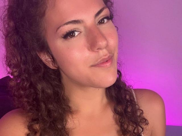 OliviaOcean-hot has curly brown hair, brown eyes, and a slight smile. She's topless with pink-purple lighting on her fair skin. Her tits are not visible.