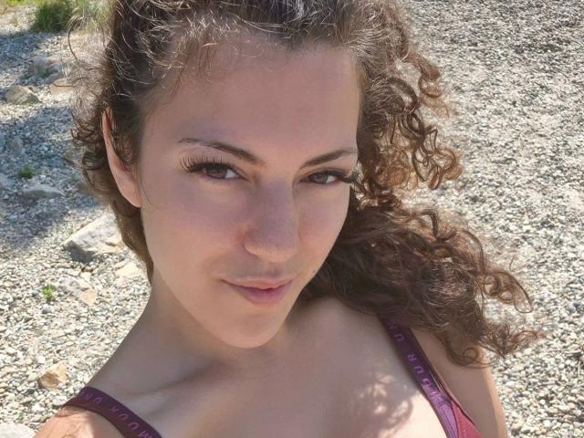 OliviaOcean-hot with curly brown hair, light skin, and brown eyes, wearing a pink bra, showing her tits, smiling slightly, in a sunny, gravelly outdoor setting.