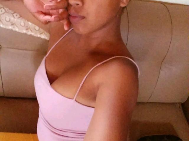 Delancie with tan skin, pink tank top, showing her tits, hand near mouth, brown sofa in background, soft light.