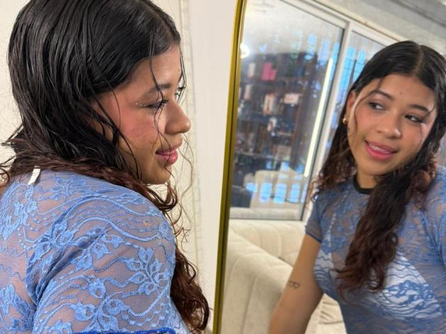IvyLancaster with wavy brown hair, wearing a blue lace top, smiling at her reflection in a mirror. Her small tits are visible through the lace.