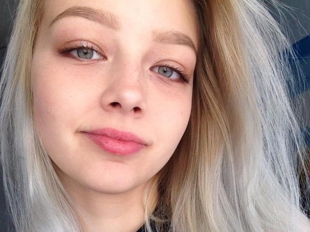 GeLik69's face, with blonde hair, light skin, pink lips, and subtle makeup, shows a slight smile. She has grey eyes, thin eyebrows, and long, straight hair.