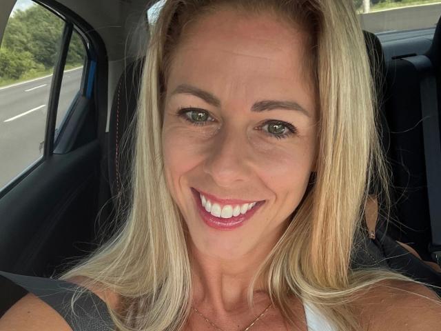 Tittney-hot tem cabelo loiro, olhos verdes e um grande sorriso com batom rosa, vestindo uma blusa branca, sentada em um carro com uma estrada visível do lado de fora..