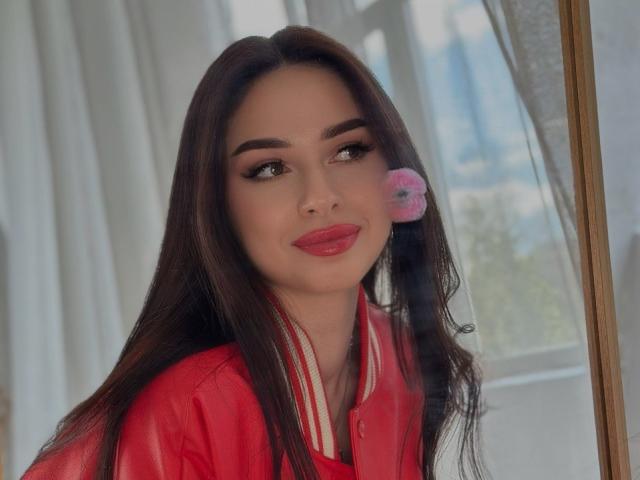 HollyJozy with long brown hair, red lipstick, and a pink flower in her hair, wearing a red shirt, looking to the side with a slight smile.