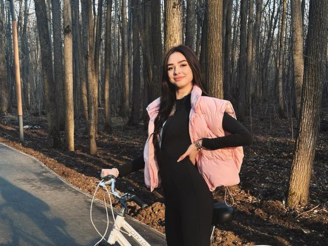 HollyJozy with long brown hair, wearing a black dress and pink puffy vest, standing by a white bike, hand on hip, in a leafless forest, smiling.