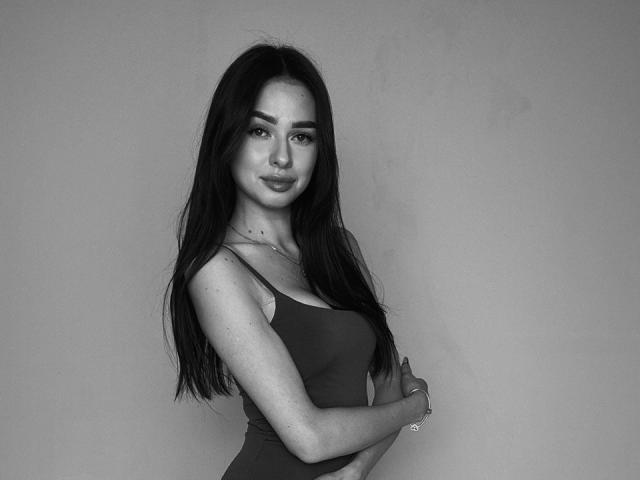 HollyJozy with long dark hair, wearing a tight tank top, her small tits and slim waist visible, standing with arms crossed, looking at the camera with a subtle smile.