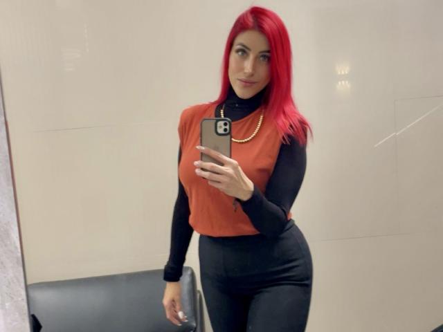 AlissaBrown with red hair, black long-sleeve shirt, orange turtleneck, black pants, holding phone, gold necklace, standing, looking at mirror, medium tits, slim, sexy.