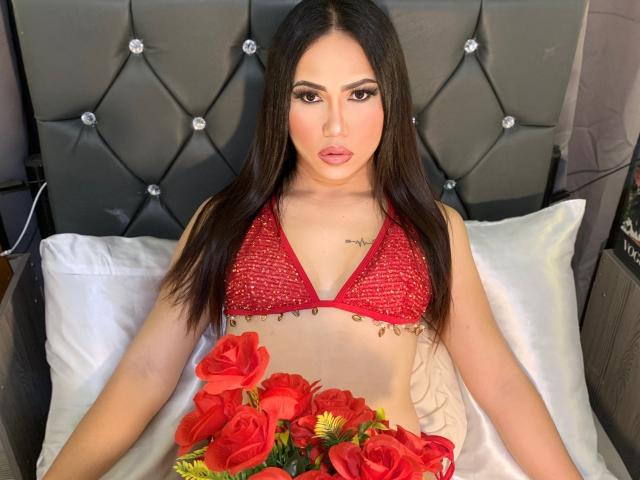 Amiraheather with long brown hair, red lace bra, and tattoo, sitting on bed with white pillows, holding red roses, looking directly at camera, sexy, cute, and attractive.