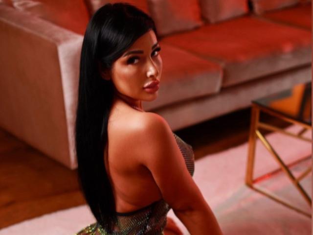 TifanyKiss has long black hair, looking over her shoulder, wearing a strapless top that shows her ass, sitting in a room with red lighting.