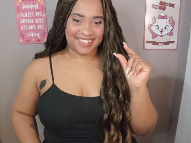 SuelenBarbar4-hot has long brown wavy hair, a black ribbed tank top, and is smiling with a hand up, showing off white nails. She has medium tits and a tattoo.