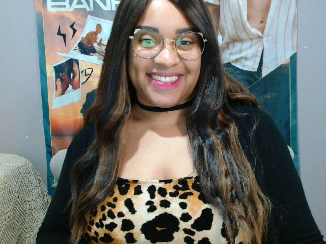 SuelenBarbar4-hot with long wavy brown hair, glasses, pink lipstick, black choker, leopard print top, black cardigan, smiling, sitting, photo showing her cute face and sexy cleavage.