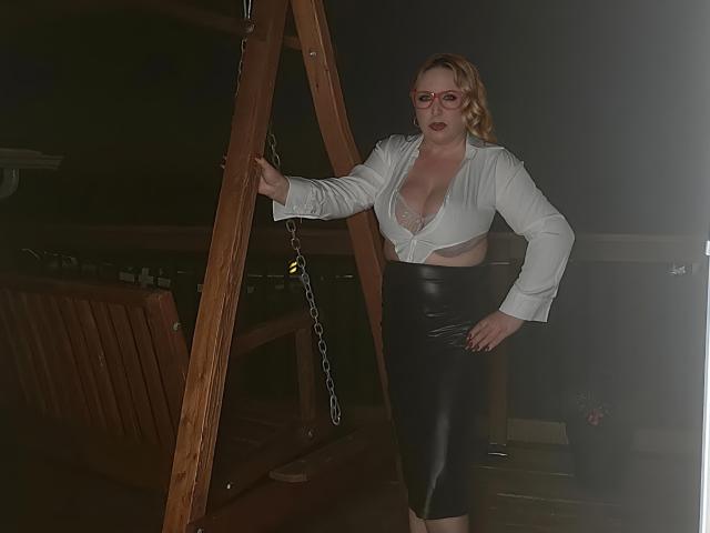 TitanicTits with blonde hair, red glasses, white tied-up shirt revealing large boobs, black leather skirt, standing by wooden structure, hand on hip, looking confident.