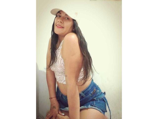 ScarletJamiesson with long black hair, white cap, white patterned top, and blue denim shorts, smiling and leaning forward, showing her small tits and slim ass.