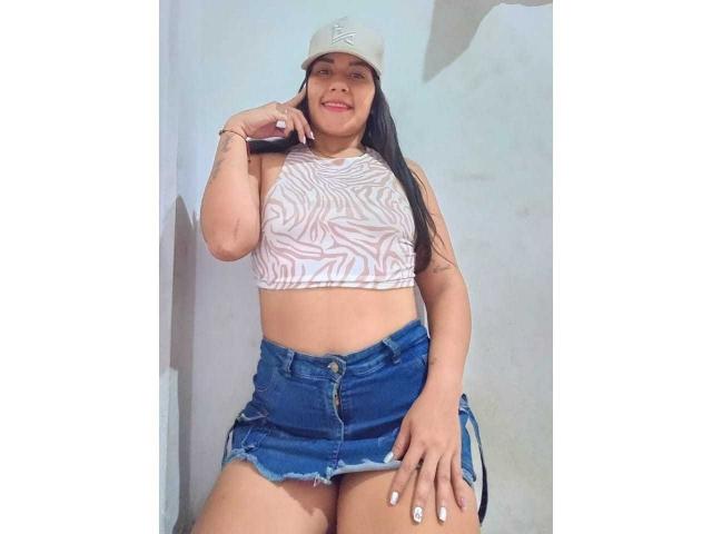 ScarletJamiesson with long black hair, wearing a white zebra-patterned crop top and blue denim skirt, smiling, hand near face, white cap, white nail polish, standing.