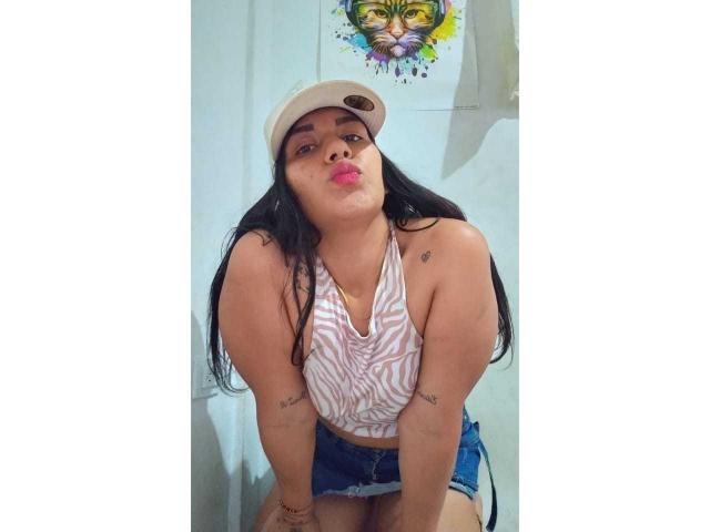 ScarletJamiesson has long black hair, wears a beige cap, pink-striped top, and denim shorts, pouting with pink lips, tattoos on arms, and a colorful face tattoo above her head.