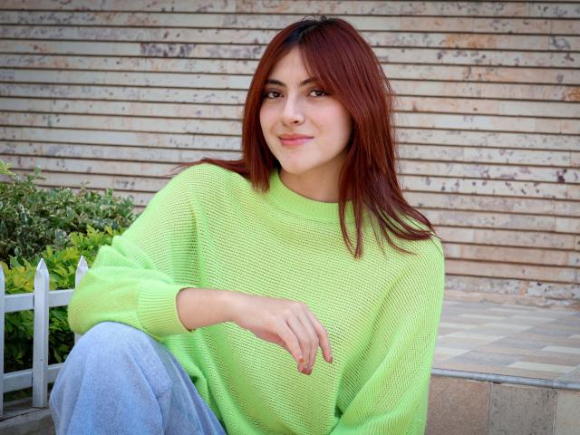 SpencerLe, con i capelli rossi, un maglione verde a maglia e jeans blu, seduta con un leggero sorriso, una mano sul ginocchio, uno sfondo di parete in legno e una pianta verde a sinistra..