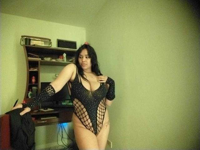 Shalyn, con grandes senos, vestida con un traje de baño negro de malla, de corte alto, que revela su vulva, está de pie en una habitación, apoyada en un escritorio, con el pelo largo y negro, y una ligera sonrisa..