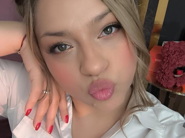 AgathaWalker has pouty pink lips, green eyes, and red nails. She's wearing a white shirt with blonde hair in a messy updo. Her ringed hand rests on her head.
