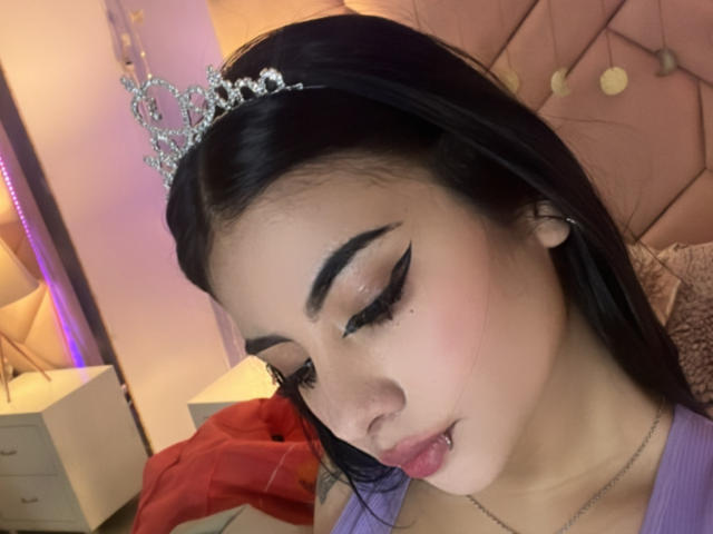 NatallieBlue with dark hair, wearing a sparkly tiara, has bold winged eyeliner, pink blush, and a silver lip piercing. She's in a purple top, looking down.