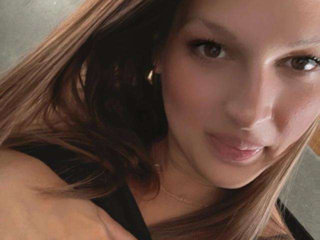 Elli69-hot with brown eyes, light skin, and straight brown hair, smiling slightly, wearing a black top, small gold hoop earrings, and a thin gold necklace.