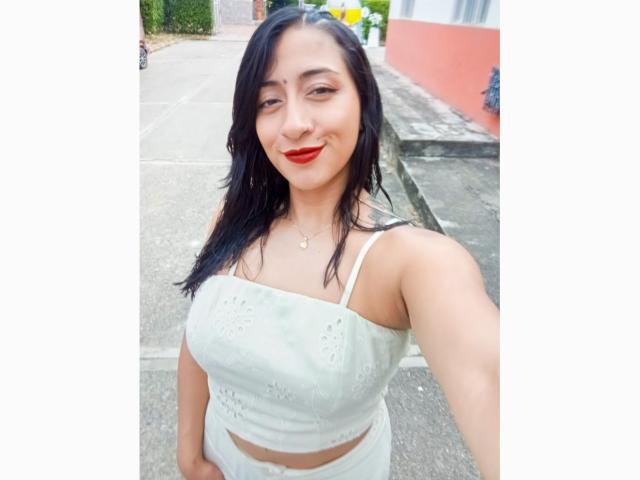 ZaraBeicker with dark hair, red lipstick, white lace crop top, showing her tits, standing on a sidewalk, smiling at the camera.