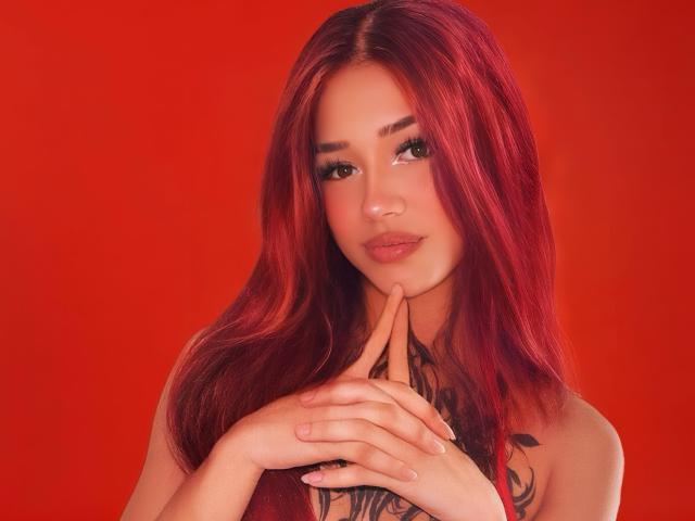 MaddyBell with red hair, holding fingers to her chin, looking at camera, tattoo on chest, red background, soft smile, slightly parted lips, cute, sexy.