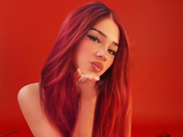 MaddyBell with long red hair, pouty lips, and thick black eyeliner, touching her chin, looking seductively at the camera. Her tits are barely visible. Red background.