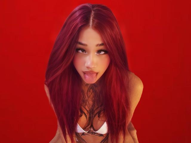 MaddyBell with red hair, sticking out her tongue, wearing a white bikini top, has tattoos on her chest, and a sexy, playful expression. Red background.