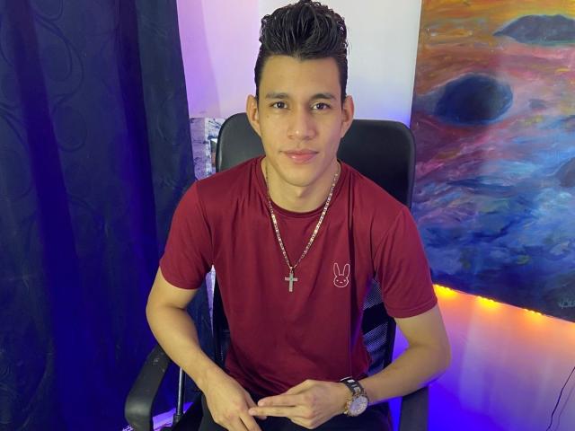 TheNewMan with short black hair, wearing a red shirt, cross necklace, and wristwatch, sitting in a black chair, pointing at his fingers, looking at the camera.