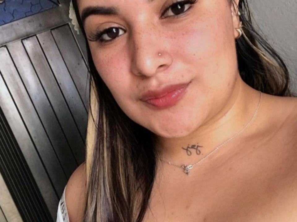 A Sanacato tem uma carinha fofa, olhos castanhos e uma pequena perfuração no nariz. Ela está sorrindo, com os seios levemente visíveis, usando um colar e com uma pequena tatuagem no pescoço..