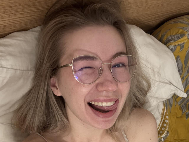 MiaFlirt with blonde hair, glasses, and a wide smile, winking, lying in bed with her tongue slightly out, showing her teeth. Her ass is partially visible.
