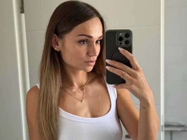 MonaMystery-hot with long brown hair, white tank top, gold necklace, and earrings, taking a selfie with a black iPhone. Boobs visible, small ring on right hand.