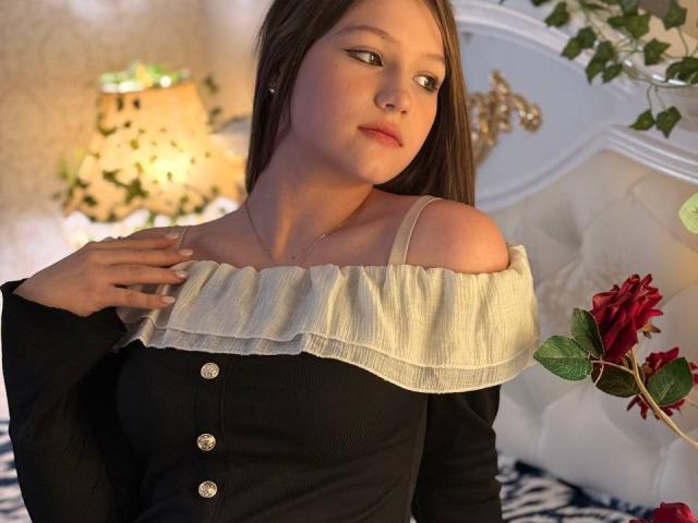 Bubochka with pale skin, long brown hair, and subtle makeup, wearing a black off-shoulder top with white ruffle, holding a red rose, in a softly lit bedroom.