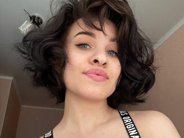 Ssimona has messy black hair, blue eyes, pink lips, and is wearing a black and white strap with "SUPER" on it. Her tits are not visible. She's smiling.
