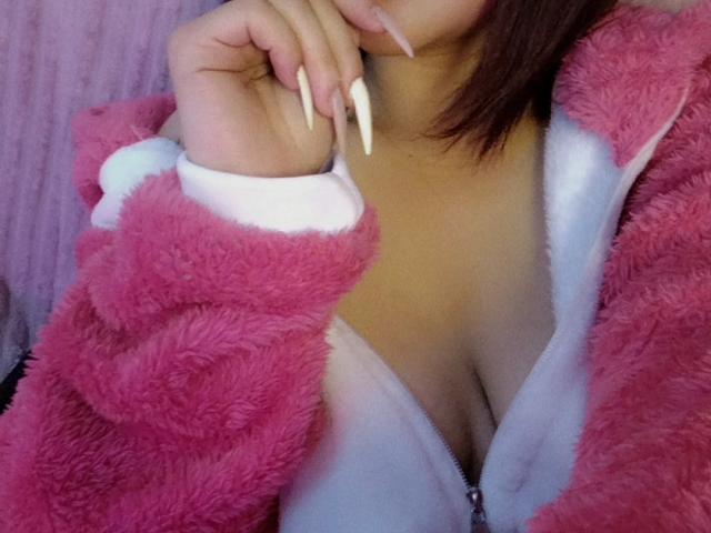 KionI's pink fuzzy jacket with white trim is open, revealing her big tits and a white zip-up top. She's biting her long, white, pointed nails.