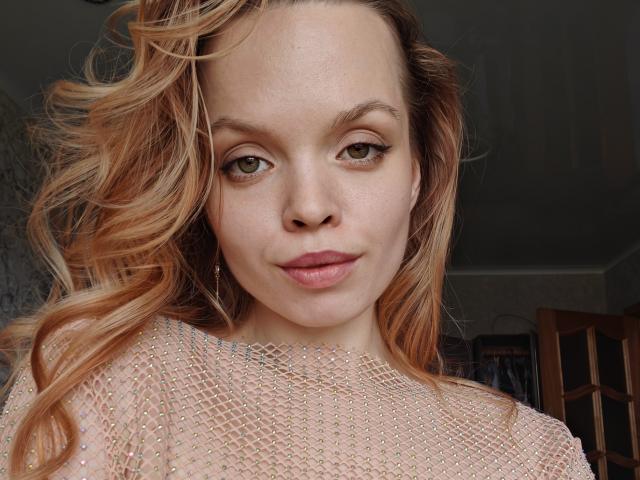 Sinteas has wavy blonde hair, green eyes, and is wearing a sheer, gold-embellished top. Her slightly parted lips and subtle smile are visible. She's looking directly at the camera.