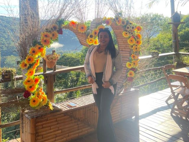 BrunaRuna with black hair, tan skin, and medium tits, wearing a beige top, black pants, and white cardigan, standing in front of a sunflower arch.