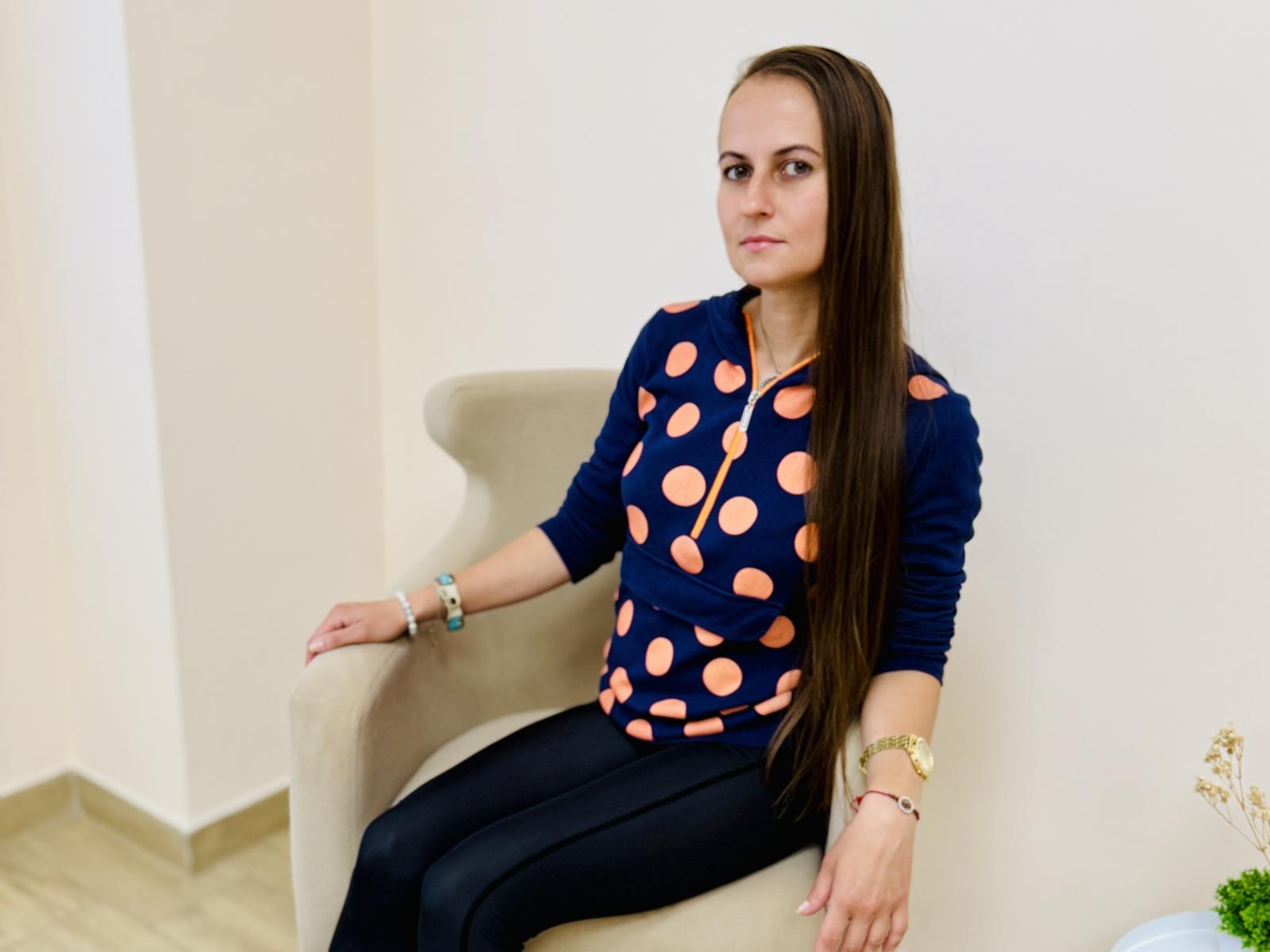Taylor Matthews, avec de longs cheveux bruns, portant une chemise bleu marine avec des pois orange, un pantalon noir et de nombreux bracelets, assise sur une chaise beige, avec une expression sérieuse..