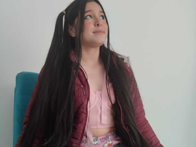 DulceTernura with long black hair in pigtails, wearing a pink bra and maroon jacket, sitting on a blue chair, looking up with a slight smile.