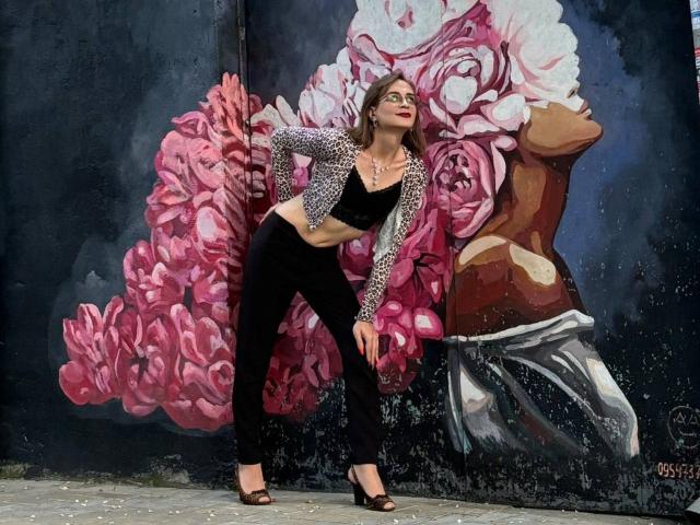 SweetyNikki stands in front of a graffiti wall, wearing a leopard print jacket, black crop top, and pants, with brown open-toe heels, leaning with a hand on her hip.
