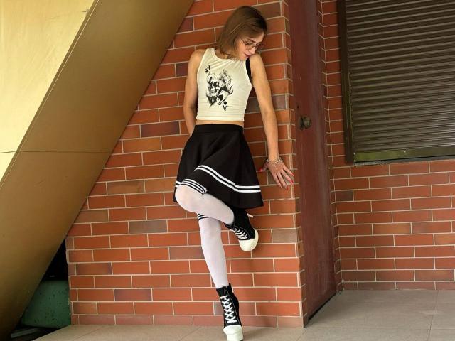 SweetyNikki has a cute ass, wearing a white tank top, black skirt, white leggings, and black/white sneakers, standing against a red brick wall, looking down, leg raised.