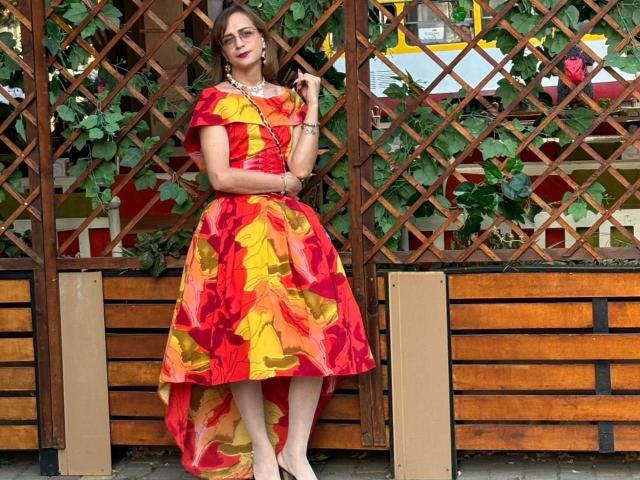 SweetyNikki with a red and yellow floral dress, holding a phone, standing against a wooden lattice, wearing a necklace and bracelets, looking cute with glasses on.