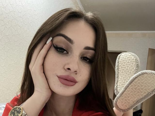 JayerSey with brown hair, red lipstick, and dramatic eyeliner, hand on cheek, holding white sneaker soles, wearing red shirt, looking at camera with slightly pouty lips.